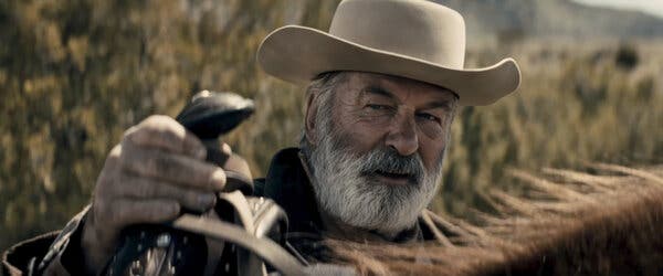 Alec Baldwin in a cowboy hat, standing behind a horse with his hand on the saddle.