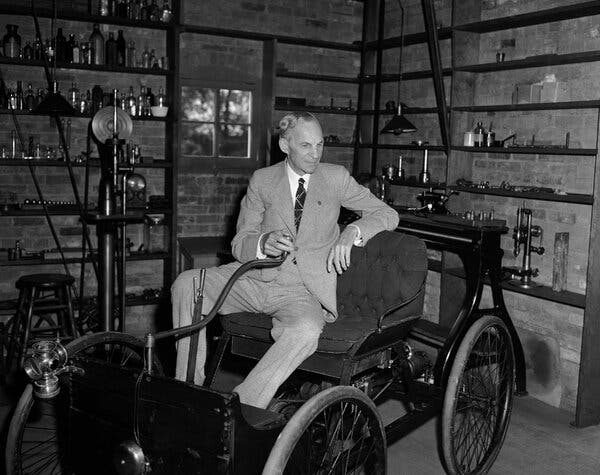 Ford is wearing a suit and sitting in an old car in a brick-walled room.